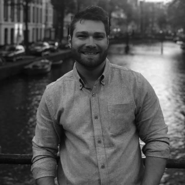 Portrait of Joe Cunningham smiling at the camera, standing outdoors on a bridge with a canal and classic European-style buildings in the blurred background, wearing a button-down shirt with rolled-up sleeves, in black and white