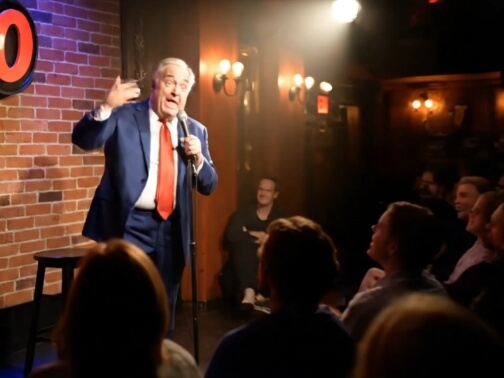An older man with white hair, wearing a blue suit, white shirt, and bright red tie, performs stand-up comedy on a dimly lit stage. He holds a microphone in one hand while gesturing expressively with the other, mouth open mid-joke. The background features a brick wall with a partial red circular sign, warm wall sconce lighting, and a small audience seated in the foreground, watching attentively under a spotlight.